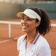 A happy woman in athletic wear smiles confidently, with a blurred tennis court background in terracotta and slate blue.